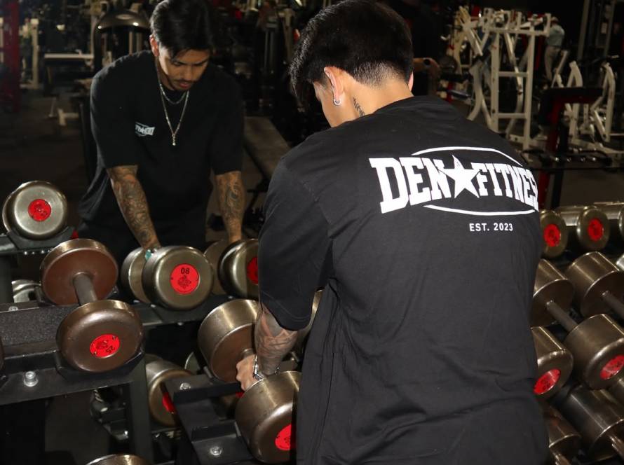 A athlete wearing a black oversized "Den Fitness EST. 2023" t-shirt seen from behind, reaching for heavy steel dumbbells on a rack in a dimly lit from a 24/7 gym, high-performance training floor.