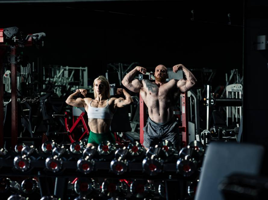 A man and a woman posing with most muscular poses in front of a mirror in a gym, surrounded by heavy dumbbells and serious training equipment.